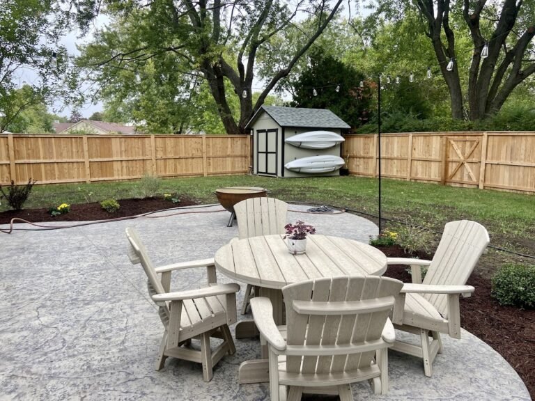 Cream table and chairs on a grey patio with a shed that has 2 kayaks hanging on it in the background.