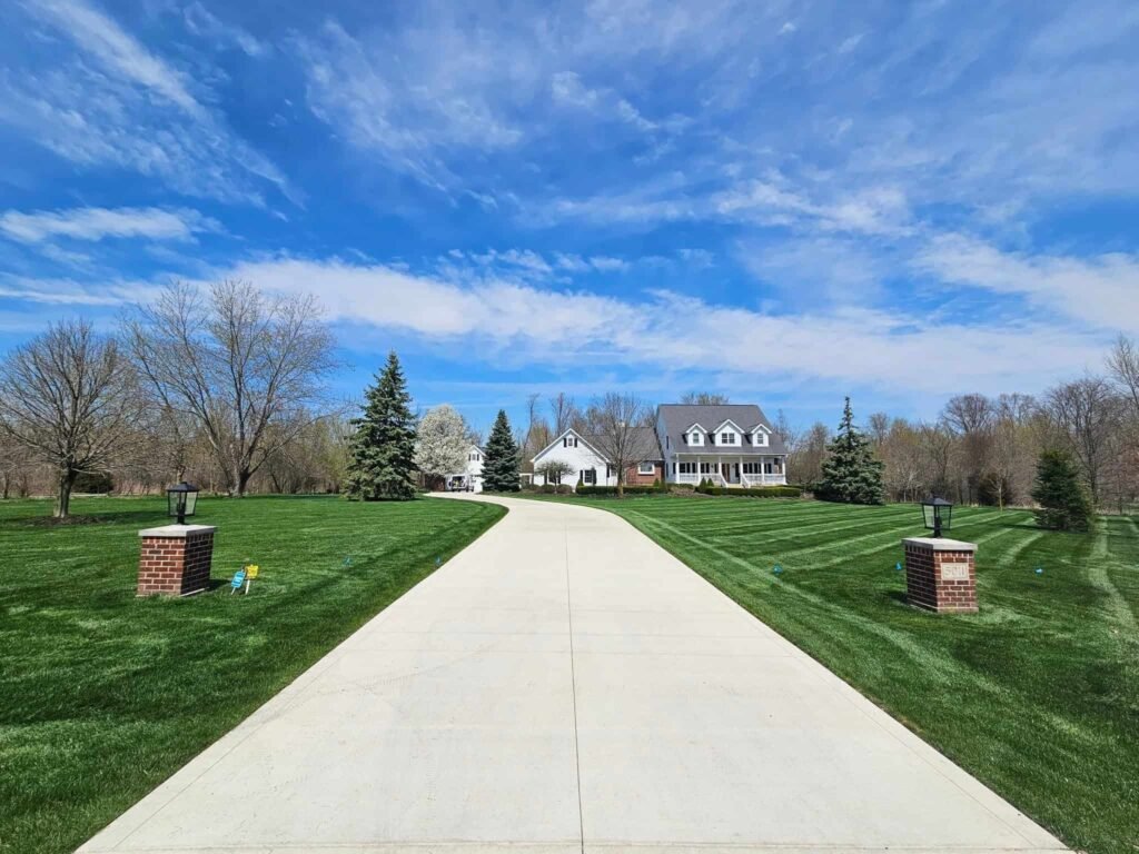 Long concrete driveway leading to a large suburban home surrounded by a beautifully manicured lawn and clear skies in Fort Wayne