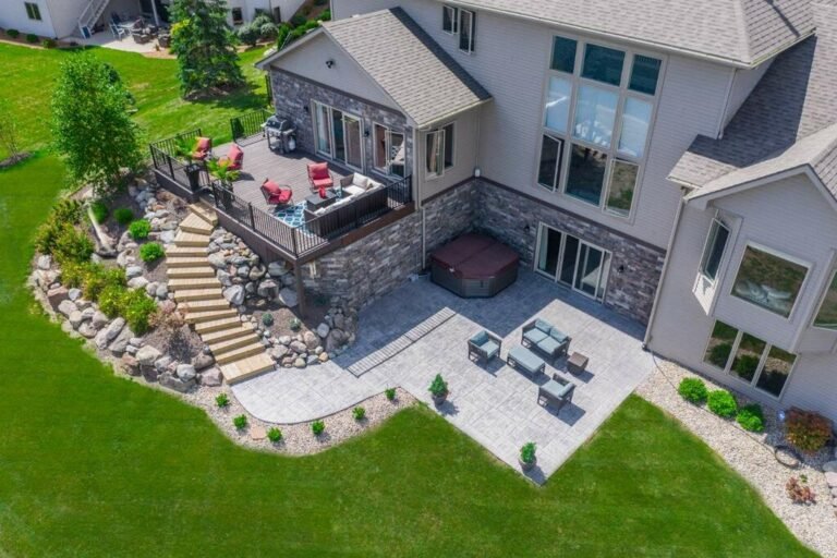 Drone shot of a grey home with stairs leading down to a patio surrounded by green grass. Showing off Crystal Creek - the best concrete contractor in Hoagland In.